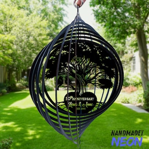 May include: Black metal wind spinner with a tree and birds design. The spinner has a spiral shape and features the text "50th Anniversary Louis & Anna EST. 1975". The background shows a green lawn and trees.