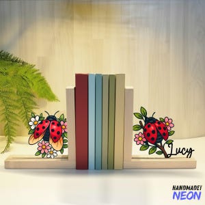 May include: Wooden bookends with ladybug and floral designs. The bookends hold a row of colorful books. One bookend has the name "Lucy" in script. The text "HANDMADET NEON" is at the bottom.