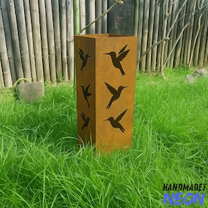 May include: A tall, rectangular, weathered metal garden planter with cut-out hummingbird silhouettes. The planter is a warm, rusty brown color and stands in a grassy area. The text "HANDMADET NEON" is visible at the bottom.
