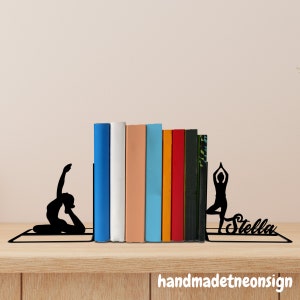 May include: Black metal bookends shaped like yoga poses, holding up a row of colorful books. The bookends are personalized with the name "Stella".