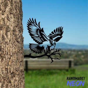 May include: Black metal silhouette of a hawk perched on a branch. The hawk is in a flying pose with its wings spread wide. The silhouette is mounted on a tree trunk.