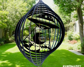 May include: A black metal wind spinner featuring a laser-cut design of a flying saucer hovering over a crescent moon and clouds. The circular object is constructed from multiple concentric rings that create a three-dimensional effect.