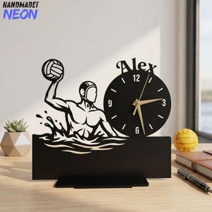 May include: A black decorative desk clock featuring a silhouette of a water polo player holding a ball above water. The circular clock face displays the name Alex at the top, with white hour markers and gold-colored hands on a solid black background.
