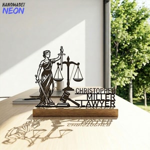 May include: A black metal sculpture of Lady Justice with a sword and scales, and the text "CHRISTOPHER MILLER LAWYER" on a wooden base. The sculpture is on a desk with a laptop and a coffee mug.