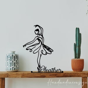 May include: Black metal silhouette of a ballerina with a flowing skirt and the name "Heather" written below. The silhouette is on a wooden shelf with a vase and a potted cactus.