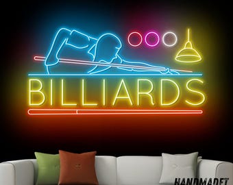 May include: A neon sign with the word "BILLIARDS" in yellow letters. The sign also features a blue silhouette of a person playing pool, with pink and purple circles representing the balls. The sign is labeled "HANDMADE NEON".