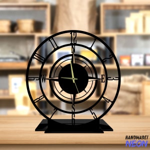 May include: Black metal clock with a compass design. The clock face has Roman numerals and a compass rose with the directions North, South, East, and West. The clock is on a black stand.