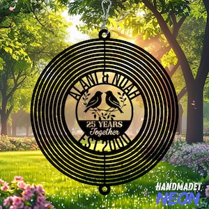 Pode incluir: Moinho de vento de metal preto com um design circular. O centro apresenta dois p&aacute;ssaros empoleirados num galho, com os nomes "ALANI & NOAH" e o texto "25 YEARS Together EST 2000". O moinho de vento &eacute; pendurado por um gancho prateado.