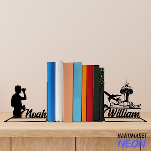 Puede incluir: Sujeta libros de metal negro con una silueta de una persona mirando a través de binoculares en el lado izquierdo y una silueta de un avión y una torre en el lado derecho. Los sujeta libros sostienen una fila de libros coloridos. Los sujeta libros están etiquetados como "Noah" y "William".