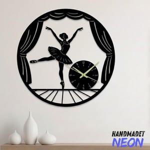 May include: Black wall clock featuring a ballerina silhouette on a stage, with a clock face at the center. The clock has a round shape and includes the text "HANDMADET NEON". The clock hands are a light green color.