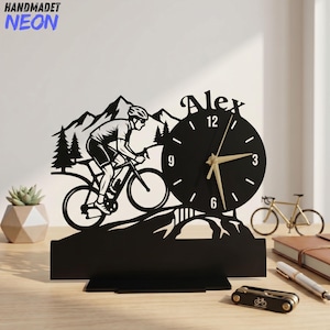 May include: A black laser-cut desk clock featuring a silhouette of a cyclist riding a mountain bike through a forest with mountains in the background. The clock face is circular with gold hands and the name Alex displayed in black lettering above it.