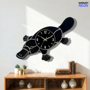 May include: A black platypus-shaped wall clock with a silver outline. The clock face is black with white numbers and hands. The clock is mounted on a white wall. The text "HANDMADET NEON" is in the upper right corner.
