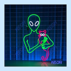 May include: Green neon alien holding a pink neon cat. The neon sign says "Handmade Neon" at the bottom.