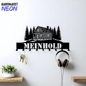 以下が含まれることがあります： 黒い金属製のキーホルダー。キャビンのデザインで、「MEINHOLD」という名前と、鍵とヘッドフォン用のフックが付いています。上部にはキャビンと木のシルエットがあります。「HANDMADET NEON」というテキストが左上にあります。
