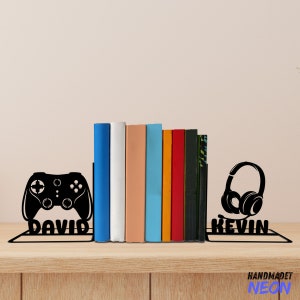 Puede incluir: Dos sujetalibros de metal negro con un diseño de mando de videojuego a la izquierda y auriculares a la derecha. Los sujetalibros sostienen una fila de libros coloridos. Los sujetalibros están etiquetados como "DAVID" y "KEVIN".
