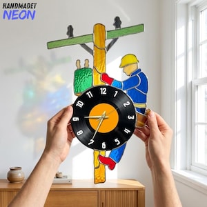 May include: A stained glass wall clock featuring a utility pole with a worker in a yellow hard hat and blue uniform. The clock face is a black vinyl record with white numbers and gold hands. The design includes green and yellow glass accents.