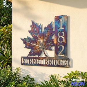 May include: A metal house number sign featuring a large maple leaf cutout next to a vertical column with the numbers 182 and a horizontal base bar with the text STREET BRIDGE. The metal has a heat-treated finish with blue and purple tones.