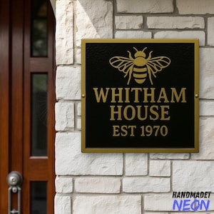 Puede incluir: Un letrero de casa negro y dorado con el texto "WHITHAM HOUSE EST 1970" y un gráfico de abeja. El letrero está montado en una pared de ladrillos blancos. El letrero tiene un borde dorado y está sujeto con tornillos dorados.