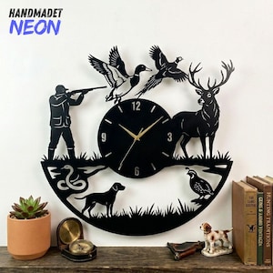 Può includere: Orologio da parete in metallo nero a tema caccia. L'orologio presenta un cervo, anatre, un cacciatore, un cane e una quaglia. Le lancette dell'orologio sono dorate. Il testo "HANDMADET NEON" è in alto a sinistra.