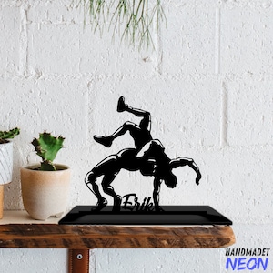 May include: Black metal silhouette of two wrestlers in a grappling pose. The silhouette is mounted on a black base with the name "Erik" written below. The base is on a wooden shelf.