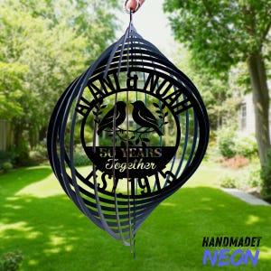 May include: Black metal wind spinner with a circular design featuring two birds and the text "BLAN & NOAH" and "50 YEARS Together EST 1974". The wind spinner is suspended outdoors against a backdrop of green grass and trees.