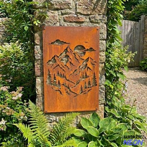 May include: A rectangular, rusty-orange metal wall art piece featuring a mountain and forest scene cut-out design. The artwork includes a sun, clouds, and trees. The piece is mounted on a stone wall and surrounded by greenery.