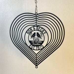 May include: Black metal heart-shaped wind spinner with concentric circles. The center circle features the names "Ron & Becky", two birds, and the text "50 Years Together EST. 1975". A decorative item for a wedding anniversary.