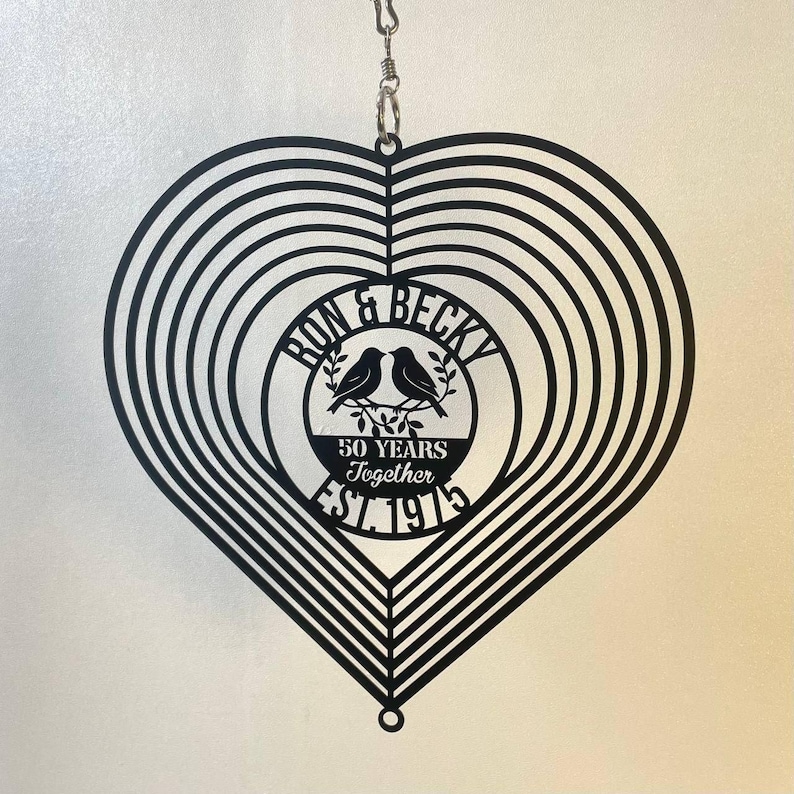 May include: Black metal heart-shaped wind spinner with concentric circles. The center circle features the names "Ron & Becky", two birds, and the text "50 Years Together EST. 1975". A decorative item for a wedding anniversary.