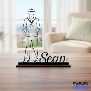 May include: A black metal sailor silhouette with the name "Sean" in cursive script. The sailor figure is wearing a hat, uniform, and has hands behind the back. The base is rectangular. The word "HANDMADET NEON" is in the bottom right corner.