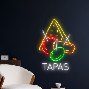 May include: Neon sign with the word "TAPAS" in white, and a yellow triangle with red and green circles and a brown rectangle inside. The sign is on a dark blue wall.