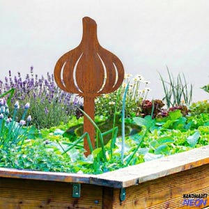 May include: A rustic, metal garlic garden stake. The stake is made of weathered, brown metal and features a detailed garlic bulb design with cut-out sections. It is placed in a raised garden bed filled with green plants and purple flowers.