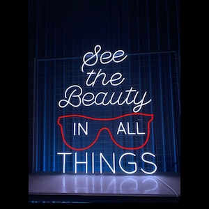 May include: A neon sign with the words "See the Beauty in All Things" in white script. A red outline of glasses is in the center. The sign is mounted on a blue grid background.