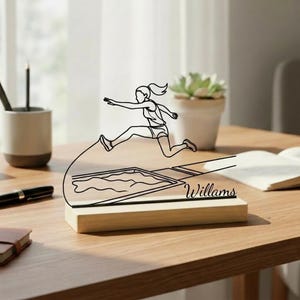 May include: A wooden base featuring a black line drawing of a long jumper in action. The name "Willams" is printed in black on the base. The drawing is placed on a wooden table, with a pen, pencil, and a small succulent plant visible.