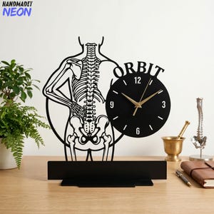 May include: Black metal wall clock featuring a skeleton design. The clock face is black with gold hands and the word "ORBIT" above. The clock is mounted on a stand with a skeleton figure.