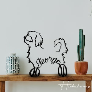 Könnte beinhalten: Schwarze Metallsilhouette eines Hundes mit dem Namen "George" darunter. Die Silhouette steht auf einem Holzregal mit einer Vase und einer Topfpflanze im Hintergrund.