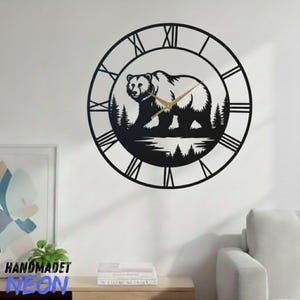 Op de afbeelding: Zwarte metalen wandklok met een berensilhouet in een boslandschap. De klok heeft Romeinse cijfers en goudkleurige wijzers. De klok is aan een witte muur bevestigd. Het woord "HANDMADET NEON" is onderaan zichtbaar.