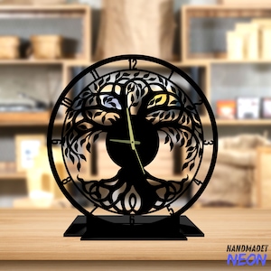 May include: Black metal clock featuring a tree of life design. The clock face is round and has Roman numeral hour markers. The clock is on a black stand.
