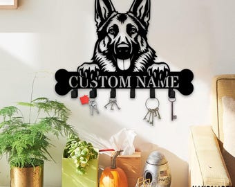 Puede incluir: Un soporte de pared para llaves de metal negro con la silueta de un perro pastor alemán apoyando sus patas sobre una barra en forma de hueso. La barra incluye el texto CUSTOM NAME y tiene seis ganchos para colgar llaves.