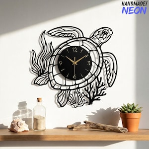 May include: A black metal wall clock shaped like a sea turtle with seaweed and coral accents. The clock face is circular with gold hands and numbers 3, 6, 9, and 12. The text HANDMADET NEON is visible in the top right corner of the image.