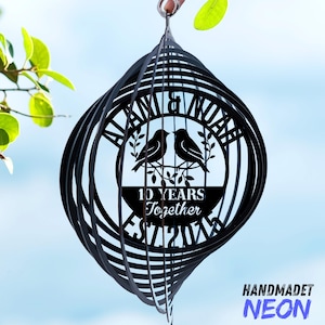 Puede incluir: Molinillo de viento de metal negro con dise&ntilde;o en espiral. El centro presenta los nombres "ALANA & NOAH" y el texto "10 YEARS Together EST 2014" con dos p&aacute;jaros posados en una rama. La palabra "HANDMADET" y la palabra "NEON" est&aacute;n en la esquina inferior derecha.