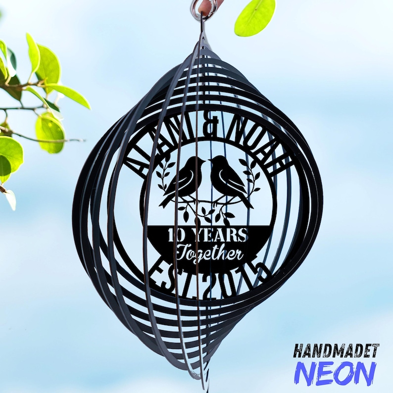 Puede incluir: Molinillo de viento de metal negro con dise&ntilde;o en espiral. El centro presenta los nombres "ALANA & NOAH" y el texto "10 YEARS Together EST 2014" con dos p&aacute;jaros posados en una rama. La palabra "HANDMADET" y la palabra "NEON" est&aacute;n en la esquina inferior derecha.