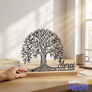 May include: A black laser-cut tree silhouette featuring intricate branches, leaves, and small birds, mounted on a rectangular light wood base. The design includes the text "Merci" above the years "2025-2026" in a black script font.