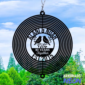 Puede incluir: Molinillo de viento de metal negro con un dise&ntilde;o circular. El centro presenta dos p&aacute;jaros posados en una rama, con los nombres "ALANI & NOAH" arriba y "10 YEARS Together EST 2015" abajo. El fondo muestra un cielo azul y &aacute;rboles verdes.