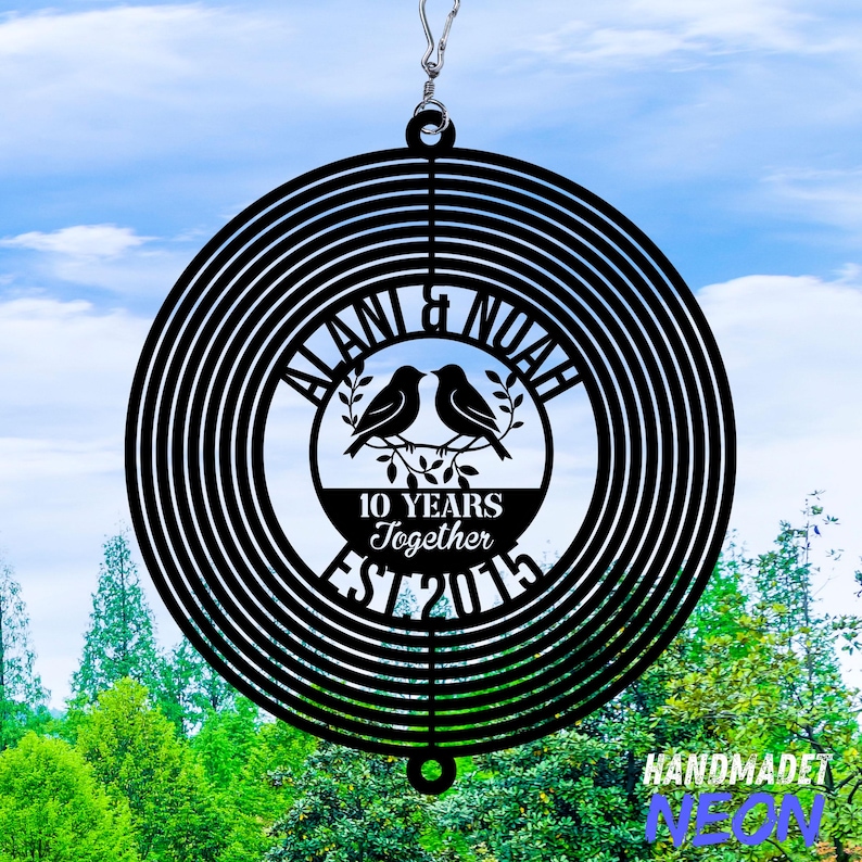 Puede incluir: Molinillo de viento de metal negro con un dise&ntilde;o circular. El centro presenta dos p&aacute;jaros posados en una rama, con los nombres "ALANI & NOAH" arriba y "10 YEARS Together EST 2015" abajo. El fondo muestra un cielo azul y &aacute;rboles verdes.