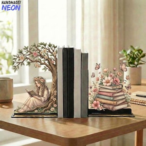 Puede incluir: Juego de sujetalibros decorativos de metal. Uno muestra a una mujer leyendo bajo un árbol, el otro una pila de libros con flores y mariposas. Los sujetalibros están sobre una mesa de madera.