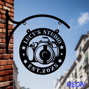May include: A black circular metal sign featuring a cutout camera graphic in the center. The text reads LUCY'S STUDIO and EST. 2025, accented by stars. The sign hangs from a curved black metal bracket attached to a brick wall structure.