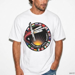 May include: A white short-sleeved t-shirt featuring a circular graphic with the text "PROJECT HAIL MARY". The design includes a central spacecraft illustration, a bright yellow sunburst, and a border composed of various international flags.
