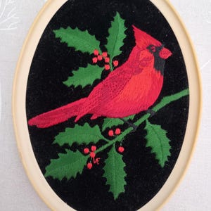 May include: Embroidered cardinal bird on a black velvet background with green holly leaves and red berries. The embroidery is framed in a cream-coloured oval frame.