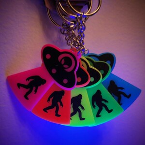 May include: Five glow-in-the-dark keychains featuring a UFO and a Bigfoot silhouette. The keychains are in a rainbow of colors: pink, orange, green, blue, and purple.