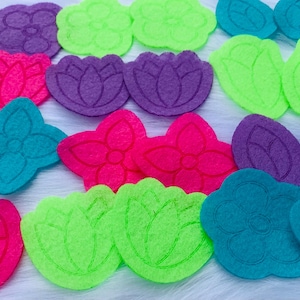 Peut inclure: Une collection de formes de fleurs et de feuilles en feutre coloré, dans diverses nuances de violet, rose, vert et sarcelle. Les formes comprennent des fleurs à quatre pétales, des fleurs de lotus et des motifs de feuilles. Ce sont probablement des fournitures d'artisanat.